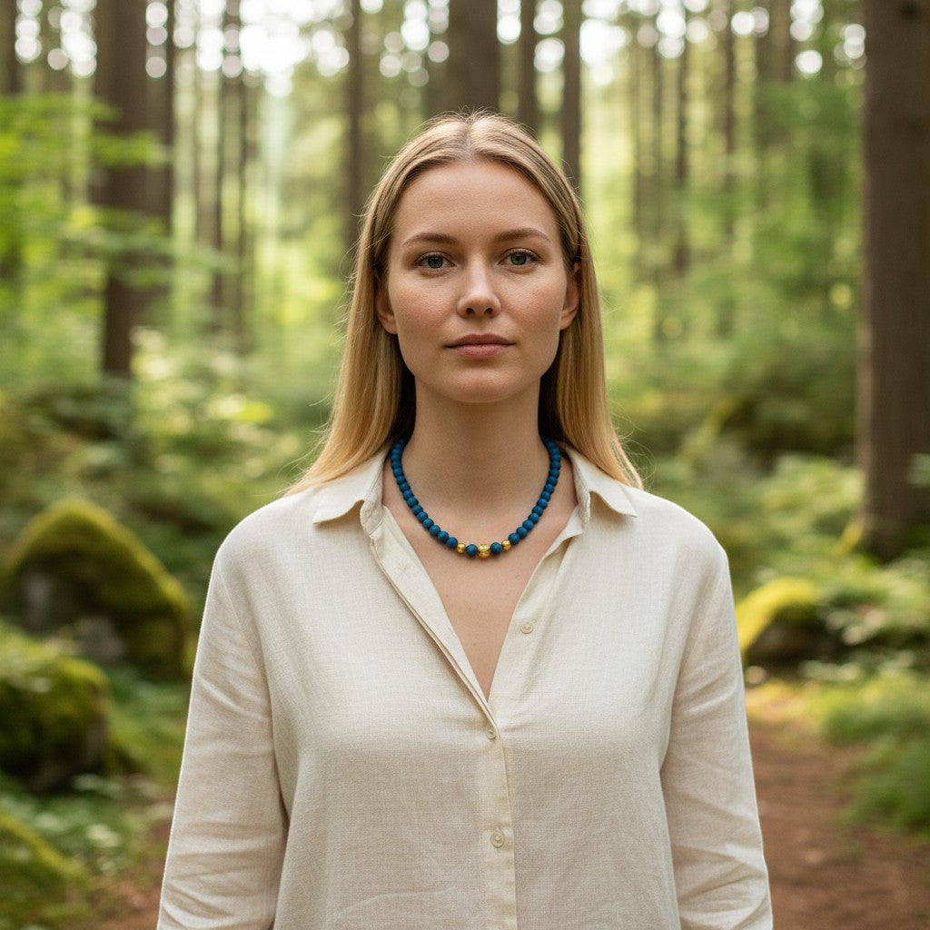 Eine Halskette aus Perlen aus blauem Achat-Geode von Sinnikäs mit goldenen Akzentperlen, getragen von einer Frau in einem Leinenhemd im Wald.