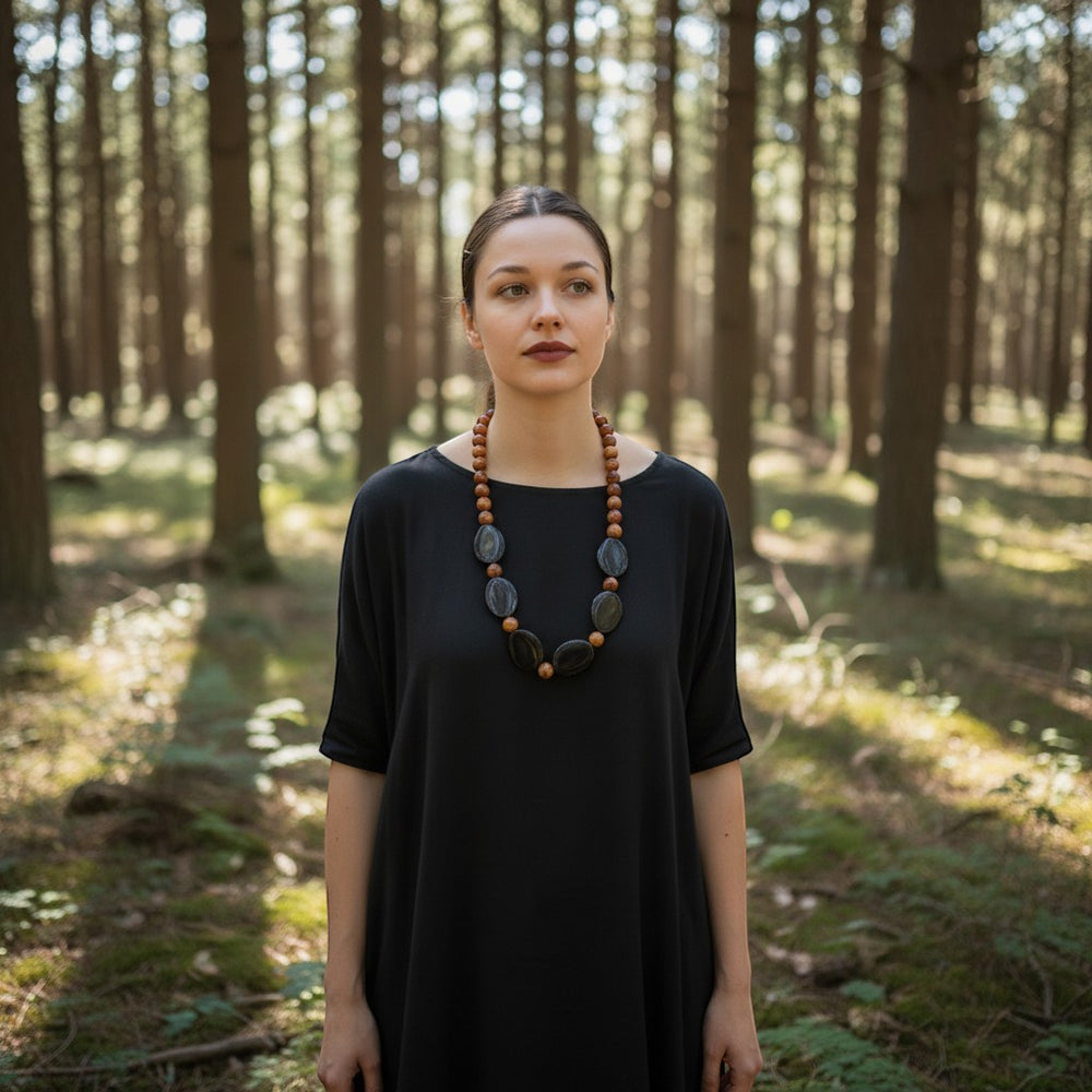 Eine auffällige Statement-Halskette mit ovalen Steinen aus tibetischem Achat und Onyx sowie runden Holzperlen. Das Model trägt ein schwarzes Kleid vor einer Waldkulisse.