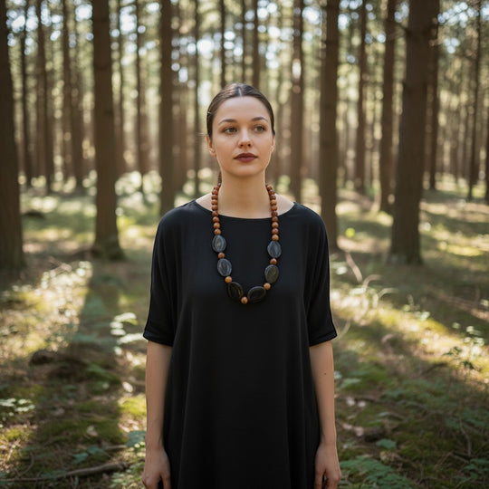 Eine auffällige Statement-Halskette mit ovalen Steinen aus tibetischem Achat und Onyx sowie runden Holzperlen. Das Model trägt ein schwarzes Kleid vor einer Waldkulisse.